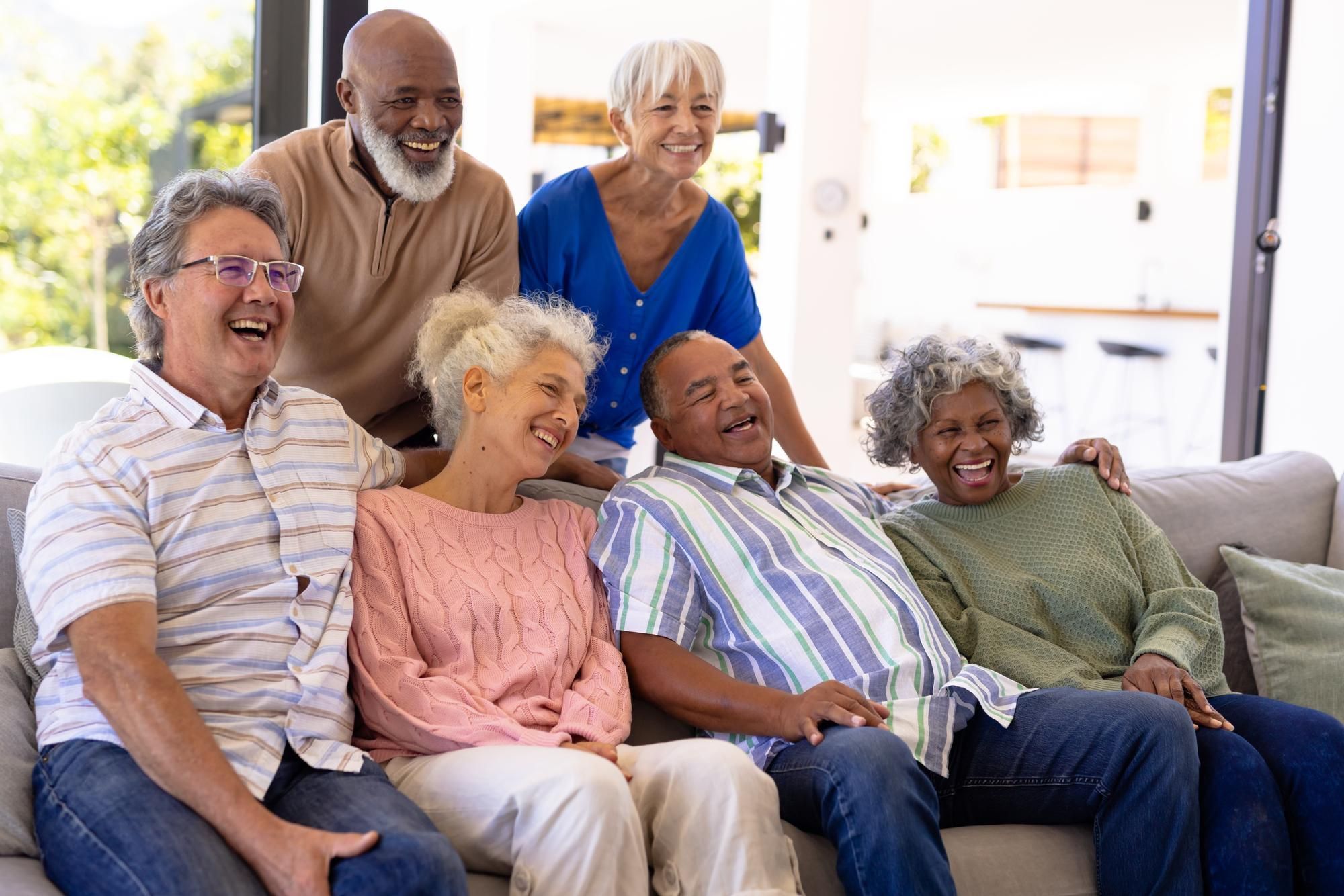 A diverse group of senior friends laughing and relaxing together on a couch in a bright, cheerful living room.