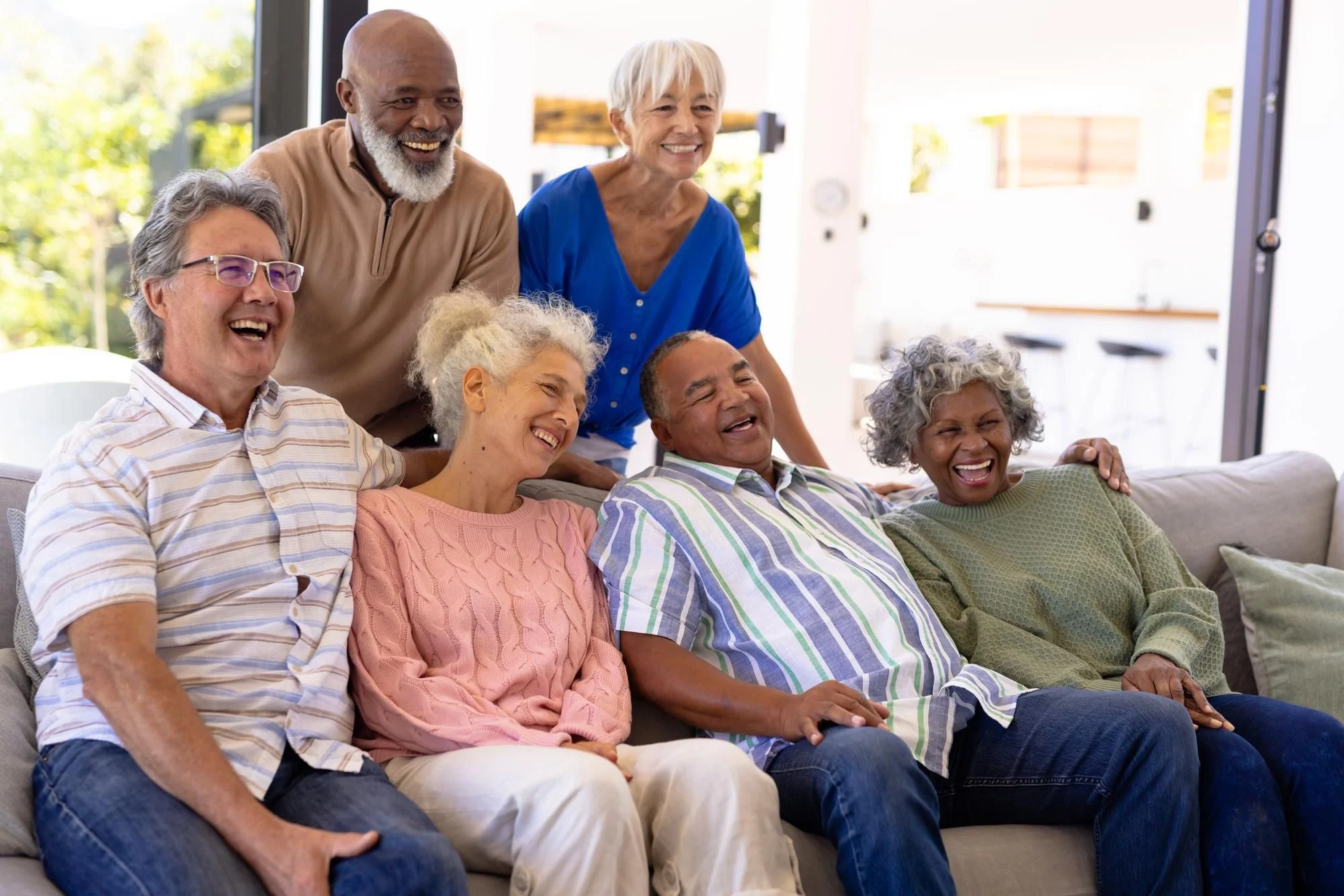 A diverse group of senior friends laughing and relaxing together on a couch in a bright, cheerful living room.